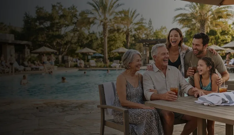 Multigenerational family laughing and enjoying cold drinks while sitting at a wooden table by a swimming pool with palm trees and lounge chairs in the background.